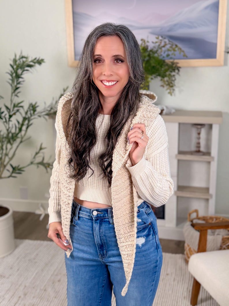 May include: A woman wearing a cream-coloured sweater and a beige crochet vest. She is also wearing blue jeans. The woman is smiling and looking at the camera. The background includes a painting and greenery.