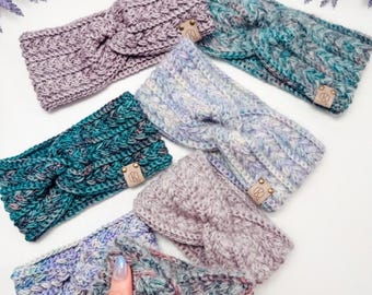 May include: A collection of seven chunky knit headbands featuring a twisted front design. Each headband is crafted from variegated yarn in shades of lavender, teal, blue, and grey, and includes a small rectangular leather tag with a logo.