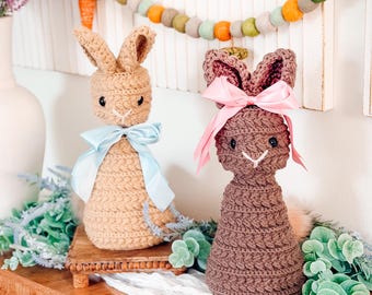 May include: Two crochet bunny ornaments. One is beige with a blue bow, the other is brown with a pink bow. They have long ears and are displayed on a wooden surface. A garland with felt balls and a carrot is in the background.