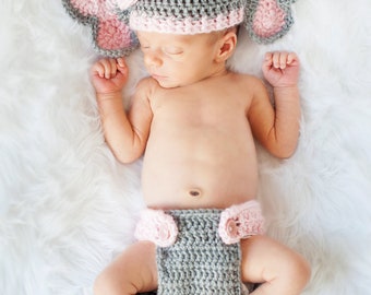 May include: A newborn baby wearing a gray and pink crocheted elephant hat and matching diaper cover. The baby is sleeping on a white fluffy blanket.