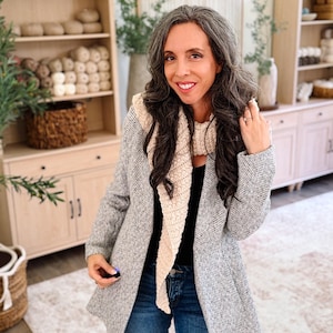 May include: A beige textured knit scarf worn over a black top and a grey herringbone blazer, paired with blue denim jeans. The scarf features a long, pointed triangular shape with a visible woven pattern, layered over the blazer and black shirt.