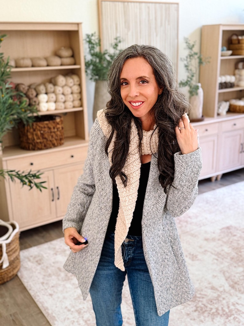 May include: A beige textured knit scarf worn over a black top and a grey herringbone blazer, paired with blue denim jeans. The scarf features a long, pointed triangular shape with a visible woven pattern, layered over the blazer and black shirt.