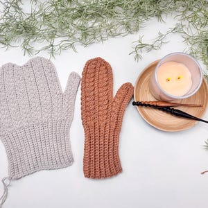 May include: Two pairs of crocheted fingerless gloves, one in a light beige colour and the other in a warm brown colour. The gloves are laid out on a white surface with a lit candle and a wooden crochet hook in the background.