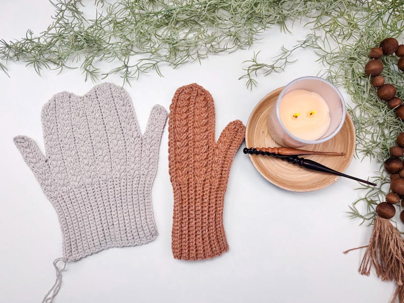 May include: Two pairs of crocheted fingerless gloves, one in a light beige colour and the other in a warm brown colour. The gloves are laid out on a white surface with a lit candle and a wooden crochet hook in the background.