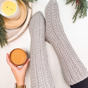 May include: A pair of gray crocheted leg warmers with a cable knit pattern. The leg warmers are worn on a person's legs, with the tops of the leg warmers folded down.