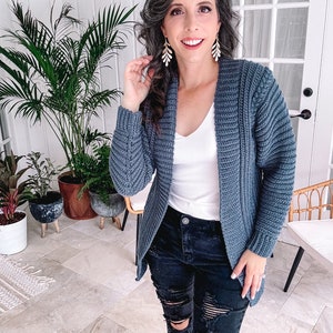 A person indoors, styled in a cochet blue cardigan, white top, and black ripped jeans, stands near plants and a chair. Their look is elevated with unique jewellery that adds a touch of elegance to their ensemble.