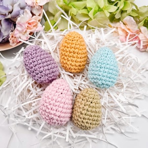 以下が含まれることがあります： 紫、黄色、青、ピンク、ベージュの色合いの、かぎ針編みで作られた5つのカラフルなイースターエッグが、白い紙のストリップの上に置かれています。 卵は花と緑に囲まれ、お祝いの春のシーンを作り出しています。