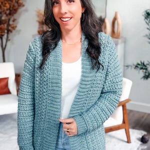 May include: A light blue crochet cardigan with a textured stitch pattern. The open-front cardigan is knee-length and has long sleeves. The woman is wearing a white top and blue jeans.