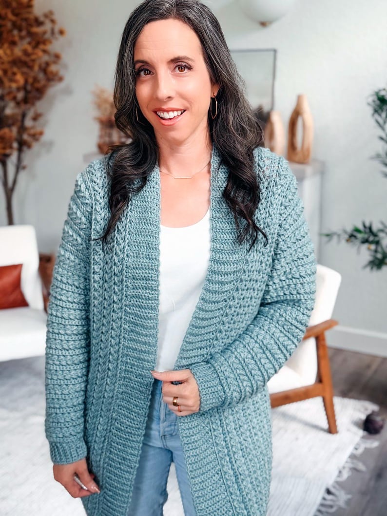 May include: A light blue crochet cardigan with a textured stitch pattern. The open-front cardigan is knee-length and has long sleeves. The woman is wearing a white top and blue jeans.