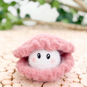 May include: A plush, pink seashell with a white, fluffy pearl inside. The pearl has black eyes and pink cheeks, giving it a cute, cartoonish appearance. The seashell is resting on a wooden surface.