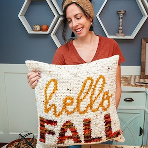 May include: A white crocheted cushion with the words "Hello Fall" in yellow yarn. The word "Fall" is in a multi-coloured stripe pattern of red, orange, and brown.