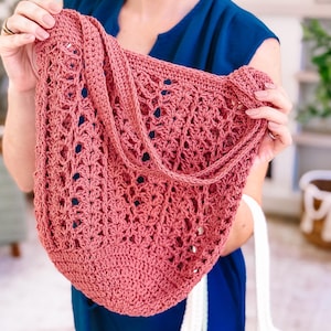 Puede incluir: Un bolso tote de ganchillo rosa polvoriento con una correa para el hombro. El bolso presenta un dise&ntilde;o abierto y calado. Una persona con una blusa azul sostiene el bolso. Un bolso de ganchillo blanco es visible debajo.