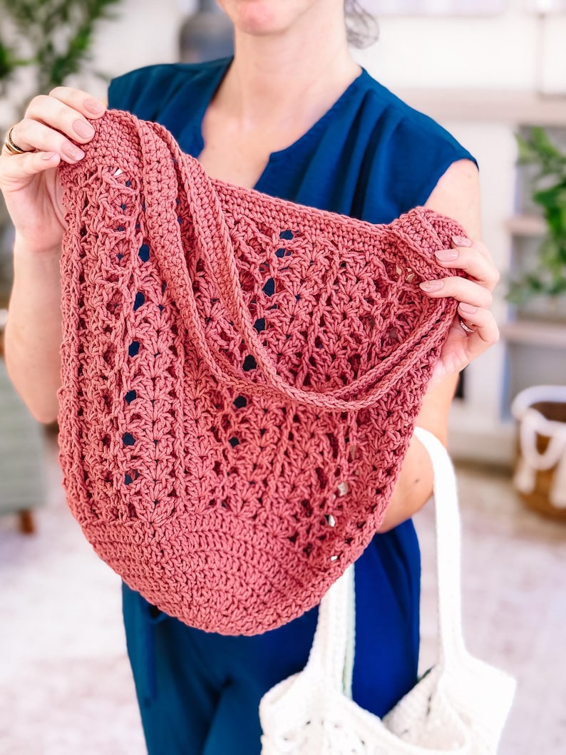 Puede incluir: Un bolso tote de ganchillo rosa polvoriento con una correa para el hombro. El bolso presenta un dise&ntilde;o abierto y calado. Una persona con una blusa azul sostiene el bolso. Un bolso de ganchillo blanco es visible debajo.