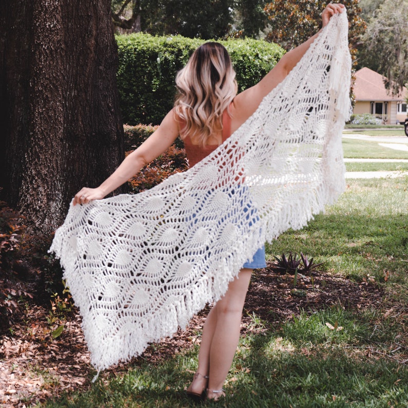 A woman wearing a white shawl decorated with the intricate crochet pineapple stitch, with fringed edges. She stands in an outdoor green space, surrounded by greenery and sunshine.