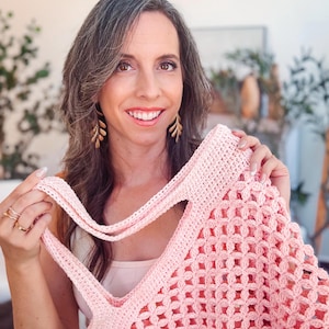 Woman with long brown hair smiles at the camera while holding up a pink crocheted tote bag—a perfect handmade gift—in a well-lit room with plants in the background.
