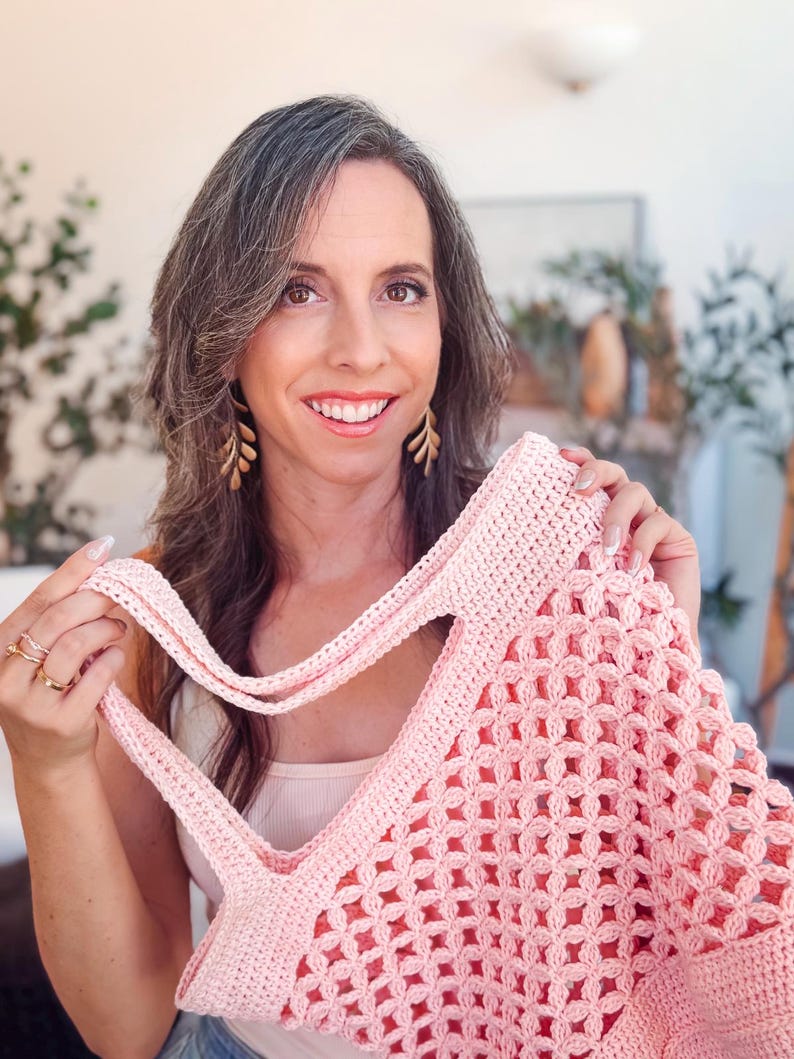 Woman with long brown hair smiles at the camera while holding up a pink crocheted tote bag—a perfect handmade gift—in a well-lit room with plants in the background.