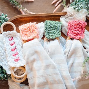 May include: A collection of handmade crochet napkin rings in various colors. The rings are made of wood and feature crochet toppers in white, pink, peach, and green. The napkins are folded and draped over a wooden tray, with floral accents.