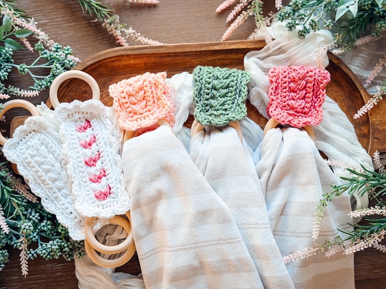 May include: A collection of handmade crochet napkin rings in various colors. The rings are made of wood and feature crochet toppers in white, pink, peach, and green. The napkins are folded and draped over a wooden tray, with floral accents.