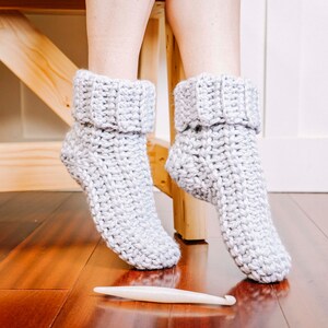 May include: A pair of grey crocheted slipper socks with a fold-over cuff. The socks are on a wooden floor with a crochet hook.