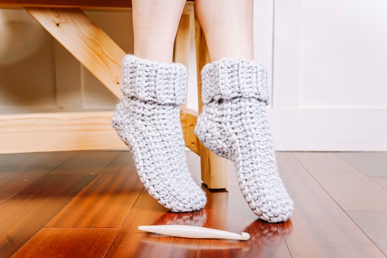 May include: A pair of grey crocheted slipper socks with a fold-over cuff. The socks are on a wooden floor with a crochet hook.