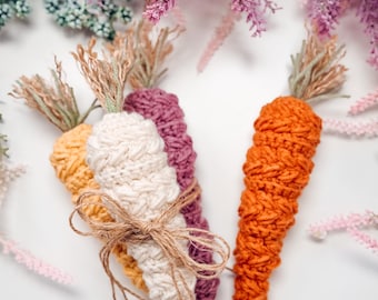 May include: Four handmade crochet carrots in orange, yellow, cream, and purple, tied with twine. The carrots have green and tan tops, and are arranged on a white surface with purple and pink floral accents.