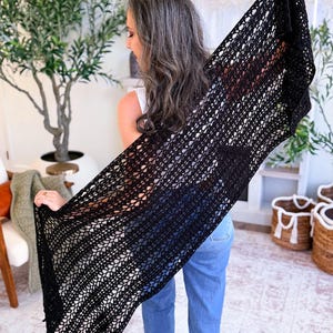 A woman with long gray hair, wearing a white top and blue jeans, holds up a large black crocheted shawl indoors—a perfect handmade gift—surrounded by plants and baskets in the background.