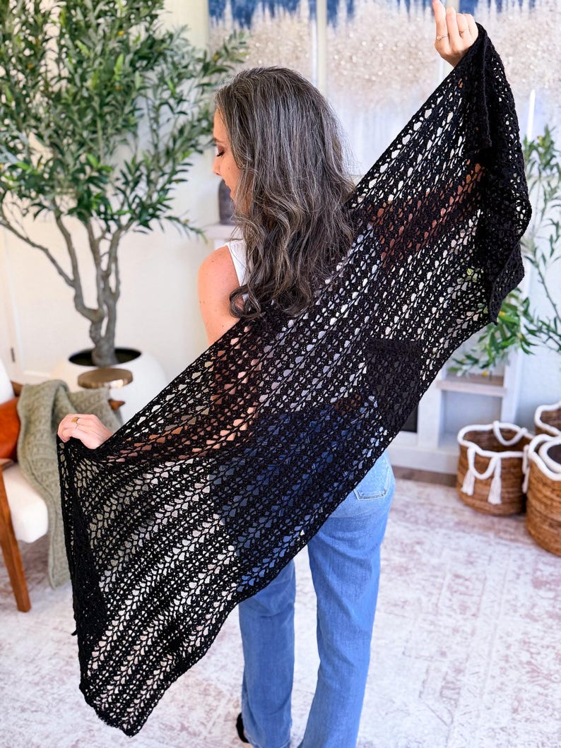 A woman with long gray hair, wearing a white top and blue jeans, holds up a large black crocheted shawl indoors—a perfect handmade gift—surrounded by plants and baskets in the background.