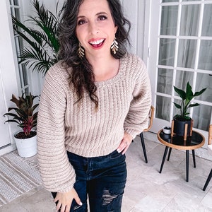 A woman with curly hair, wearing a crochet beige sweater and ripped jeans, smiles brightly indoors near potted plants, showcasing her unique jewelry.