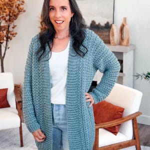 May include: A light blue, open-front cardigan with a textured knit design. The long-sleeved sweater is worn over a white top and light blue jeans. The cardigan is knee-length and appears to be handmade.