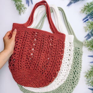 Puede incluir: Tres bolsas de mano de ganchillo en tonos rojo, blanco y verde. Las bolsas tienen asas largas y un dise&ntilde;o calado. Una mano sostiene la bolsa roja. Las bolsas est&aacute;n dispuestas sobre una superficie blanca con detalles florales.