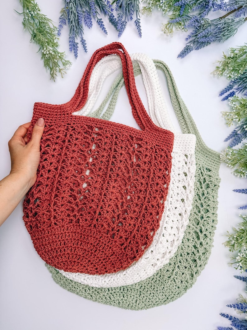 Puede incluir: Tres bolsas de mano de ganchillo en tonos rojo, blanco y verde. Las bolsas tienen asas largas y un dise&ntilde;o calado. Una mano sostiene la bolsa roja. Las bolsas est&aacute;n dispuestas sobre una superficie blanca con detalles florales.