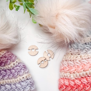 Two crocheted hats with faux fur pom-poms, one in purple tones and one in pink tones, are displayed with three wooden buttons featuring heart cutouts. Green leaves frame the top—an ideal handmade gift for any occasion.