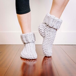 May include: A pair of grey crocheted slipper socks with a folded cuff. The socks are being worn on a person's feet.