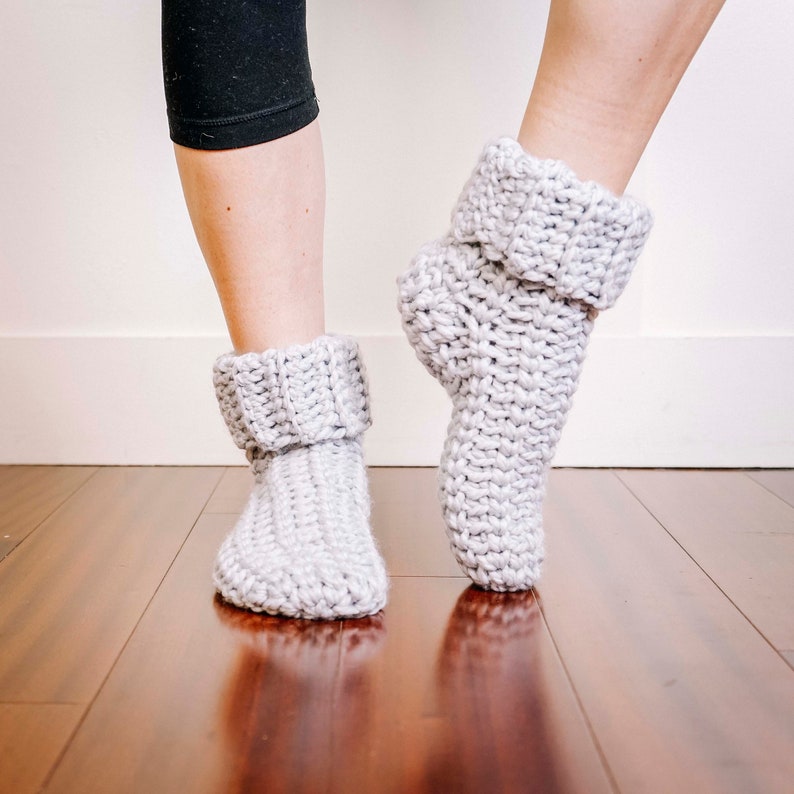 May include: A pair of grey crocheted slipper socks with a folded cuff. The socks are being worn on a person's feet.