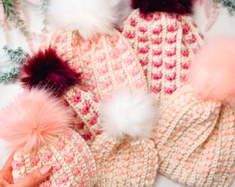 May include: A collection of handmade knitted hats in various shades of pink and cream, each topped with a fluffy faux fur pom-pom. The hats feature a textured stitch pattern and are arranged on a white surface with floral accents.