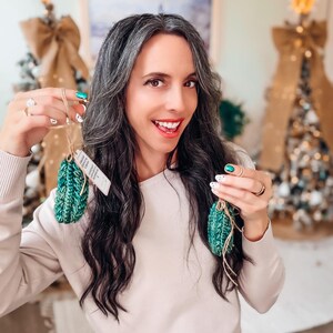 May include: Two small, textured, dark green crocheted ornaments shaped like pinecones. Each ornament features a vertical ribbed pattern and is attached to a thin, light brown twine loop for hanging. A small white paper tag is attached to one ornament.