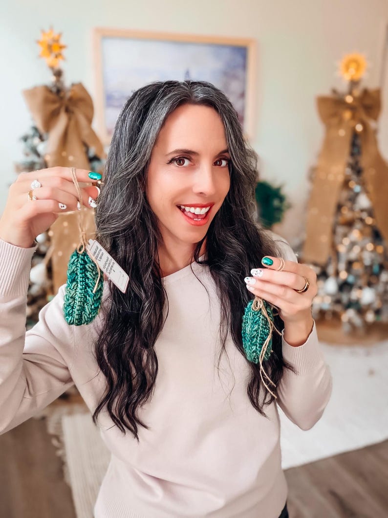 May include: Two small, textured, dark green crocheted ornaments shaped like pinecones. Each ornament features a vertical ribbed pattern and is attached to a thin, light brown twine loop for hanging. A small white paper tag is attached to one ornament.