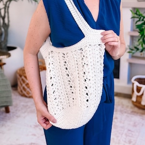 Puede incluir: Bolso tote de crochet de color crema y tejido abierto, con una correa ancha. El bolso lo sostiene una persona con un mono azul. El bolso tote tiene un fondo redondeado y un dise&ntilde;o calado texturizado, perfecto para llevar la compra.