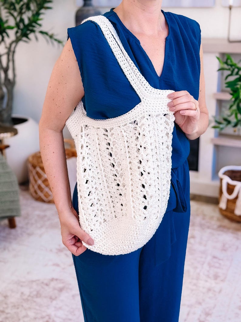 Puede incluir: Bolso tote de crochet de color crema y tejido abierto, con una correa ancha. El bolso lo sostiene una persona con un mono azul. El bolso tote tiene un fondo redondeado y un dise&ntilde;o calado texturizado, perfecto para llevar la compra.