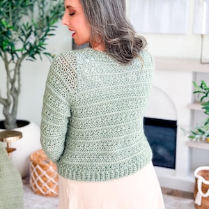 May include: A light green crochet cardigan with long sleeves. The back view shows the intricate stitch pattern and the cropped length of the sweater. The cardigan is paired with a light pink skirt. The overall style is casual and handmade.