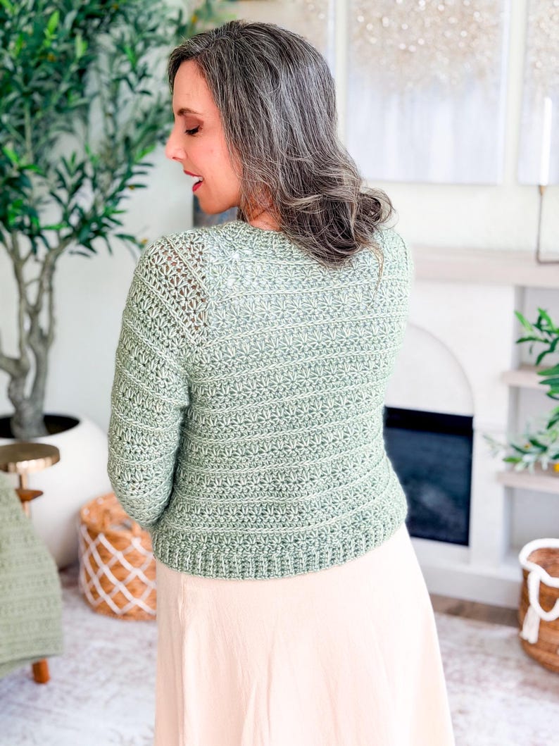 May include: A light green crochet cardigan with long sleeves. The back view shows the intricate stitch pattern and the cropped length of the sweater. The cardigan is paired with a light pink skirt. The overall style is casual and handmade.