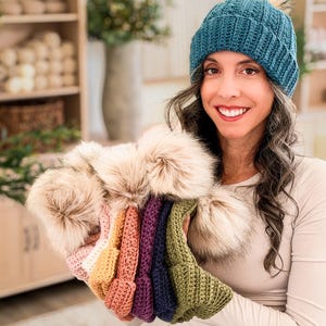 Woman in a teal crochet beanie holds a row of pom pom beanies in pink, orange, purple, navy and green