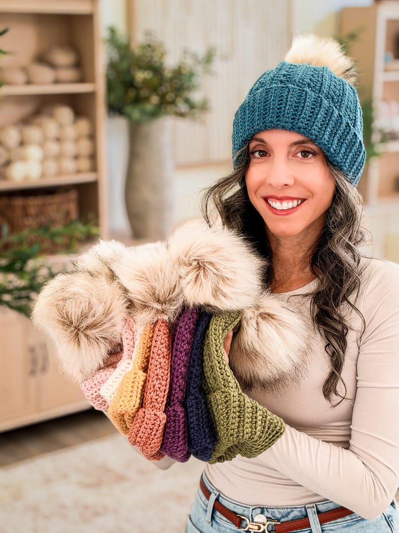 Woman in a teal crochet beanie holds a row of pom pom beanies in pink, orange, purple, navy and green