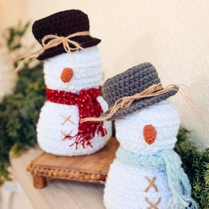 Two white crochet snowmen on a shelf, one with brown hat and red scarf, one with gray hat and blue scarf