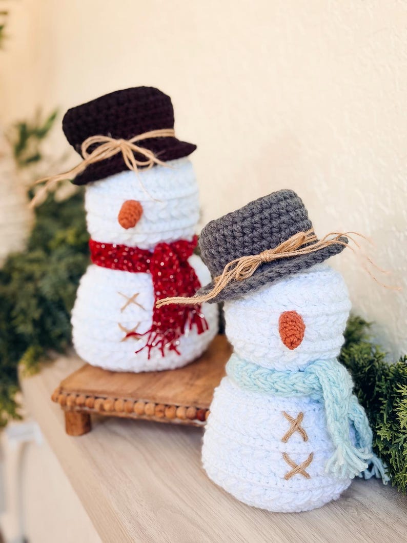 Two white crochet snowmen on a shelf, one with brown hat and red scarf, one with gray hat and blue scarf