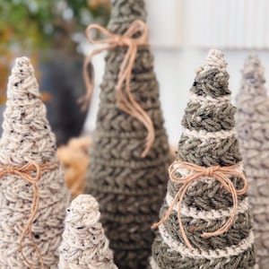 Puede incluir: Una colección de árboles decorativos en forma de cono hechos de hilo texturizado en tonos verde oliva, crema y beige moteado. Cada árbol presenta un patrón de punto de ganchillo distinto y está acentuado con un lazo de cuerda beige.