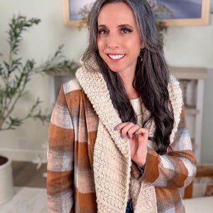 May include: A woman wearing a beige and brown checked jacket with a cream-coloured, textured scarf. The jacket has a classic checked pattern. The scarf is a lighter shade and appears to be knitted or crocheted. The woman is smiling.