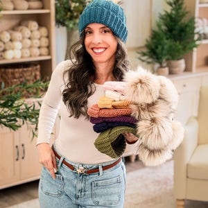 Woman in a teal crochet beanie and jeans holds a stack of colorful pom pom beanies in a cozy room
