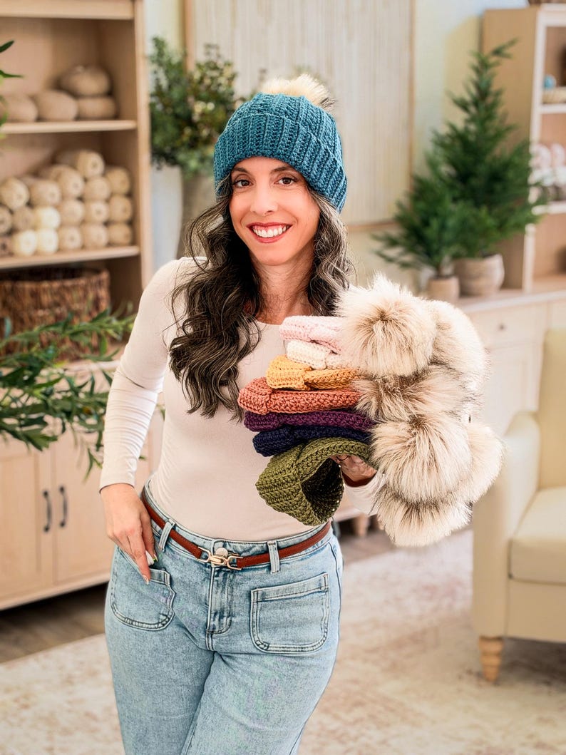 Woman in a teal crochet beanie and jeans holds a stack of colorful pom pom beanies in a cozy room