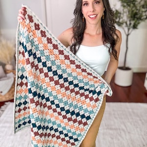 May include: A rectangular crochet blanket featuring a repeating granny square pattern in a color palette of cream, dark teal, muted orange, and deep burgundy. The blanket has a cream-colored border and a small brown rectangular tag at the corner.